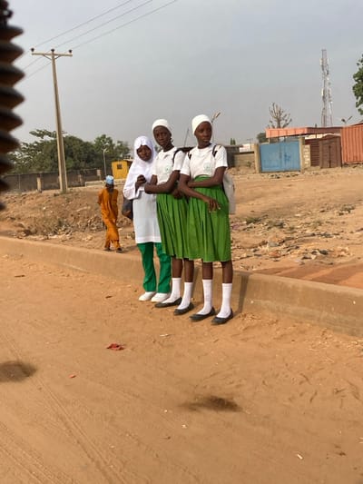 Four girls stand shoulder to shoulder on a dirt road, posing for a photo. The girls are wearing school uniforms with knee-high socks and black shoes. One girl wears a white head covering and a white tunic. Two girls wear green skirts. An adult man in a brown robe stands apart from the group. A low wall and scrubby landscape are in the background. Powerlines and a cell tower are visible in the distant sky.