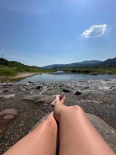 The image shows a person's legs and feet resting on a large rock in a shallow river. The water is clear and has a rocky bed. In the background, there is a sandy riverbank with grass and trees, followed by rolling hills and mountains under a bright blue sky with a single white cloud. The scene appears to be a natural, outdoor setting. The location is identified as Buk-myeon, South Korea.