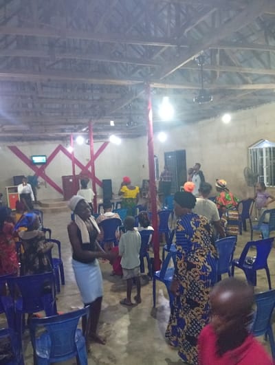 Inside a church with blue chairs and a wooden ceiling, people gather. A woman in a dark top and light skirt stands barefoot, while another wears a blue floral dress. Children are scattered among the adults. A stage decorated with red shapes is visible in the background, and a speaker system stands nearby. The room is illuminated by several bright, bare bulbs hanging from the ceiling.