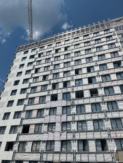 The image shows a partially completed high-rise building under construction in Аснежыцкі сельскі Савет, Belarus. A construction crane is visible in the upper left corner, with its boom extending over the building. The building's facade is a mix of white panels with a perforated pattern on the left side and exposed insulation and window frames on the right side. The insulation is covered in white plastic sheeting, with some sections revealing insulation material underneath. Numerous dark-framed windows are visible, some with plastic coverings. Metal framing for the facade is also apparent. The sky above is blue with scattered white clouds. The scene depicts an active construction site.