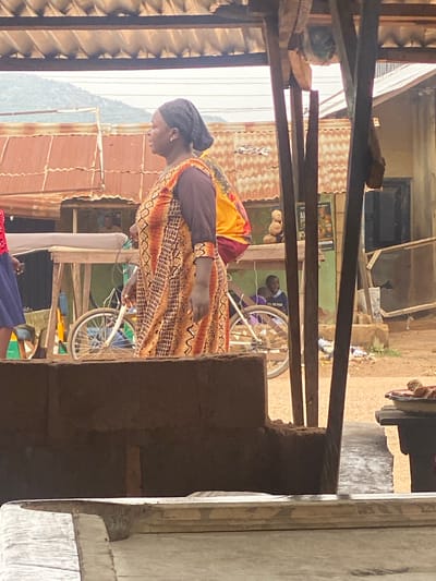 The image shows a vertical view of an outdoor scene in Barakin, Nigeria. In the foreground, a woman wearing a patterned dress and a dark headscarf is partially visible, facing away from the camera and looking down. She has on a dark brown top underneath her dress and an earring in her visible ear.
Behind the woman, the scene unfolds in what appears to be a market or a bustling outdoor area. A man and a child are near a bicycle with a white frame and spoked wheels. The man is looking towards the child, who is partially obscured. Further in the background, there are various structures, including corrugated metal roofs and wooden supports, suggesting a built environment. There are also piles of what appear to be coconuts or similar produce.
The lighting suggests it is daytime, possibly late morning or early afternoon, with sunlight casting shadows. The overall atmosphere seems informal and everyday, typical of a local community gathering or marketplace. The background shows more of the built environment, with some hints of greenery in the distance and corrugated metal structures that are common in this region. There is no visible text in the image.