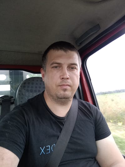 A medium shot shows a man with short brown hair and stubble sitting in the driver's seat of a vehicle, looking towards the camera. He is wearing a black t-shirt with a light blue logo on the left side of his chest that appears to read "DEX". A black seatbelt is diagonally across his chest. The interior of the vehicle is visible, including a gray headliner, a dark gray grab handle on the right, and a patterned seat behind the driver. The exterior scene through the window shows a blurred landscape of fields and trees under an overcast sky. The man is positioned in the foreground of the image, with the vehicle interior and exterior providing the background.