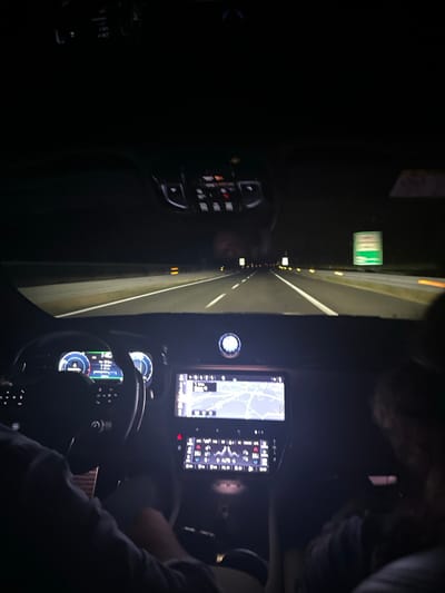 Inside a vehicle at night, illuminated displays showcase a digital map and car metrics. A steering wheel and dashboard are visible. Outside, a highway stretches ahead, marked by white lines and illuminated by intermittent lights. A green sign is seen on the roadside. The scene is dark, with the car's interior lights providing the main source of illumination.