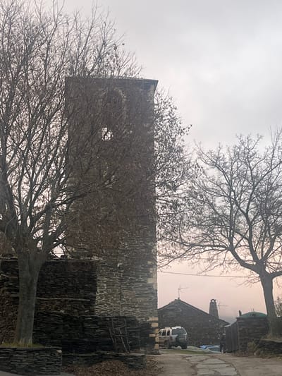 An outdoor scene in Campillo De Ranas, Spain, features prominent stone buildings and bare trees under an overcast sky. A tall, rectangular building, possibly a tower or a large house, stands in the center-left. It is constructed from dark, irregular-shaped stones, characteristic of the region's "black architecture," and has a single, high circular opening on one side. This structure is heavily framed and partially obscured by the intricate, leafless branches of several deciduous trees in the foreground and midground.

To the right, further in the background, another stone building with a dark roof, a chimney, and a television antenna is visible. A white utility vehicle, resembling an ATV or a small pickup truck, is parked beside this background building. A narrow, unpaved path or road runs along the right side of the frame, bordered by low stone walls or stacked slate. The sky is uniformly hazy and gray, indicating an overcast day, and the barren trees suggest a late autumn or winter season. Scattered fallen leaves are visible on the ground. The scene is devoid of people and active events, conveying a quiet and still atmosphere. No visible text is present.