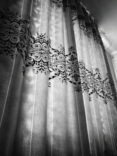 The image displays a close-up view of a translucent curtain with a black, ornate damask pattern. The curtain is draped, creating vertical pleats and folds that are accentuated by the light filtering through it. The pattern consists of swirling vines, leaves, and floral motifs, creating a decorative and somewhat vintage aesthetic. The lighting suggests it is daytime, with light coming from the top and to the right, casting subtle shadows and highlights on the fabric. The texture of the curtain appears to be fine and semi-sheer, allowing light to pass through. The scene is indoors, likely a room, given the presence of a ceiling and what appears to be molding at the top right. There are no people visible in the image. The overall mood is calm and slightly decorative due to the intricate pattern on the curtain. The location context provided is Баяндай, Kazakhstan, but there are no visual cues in the image itself to confirm this specific location.