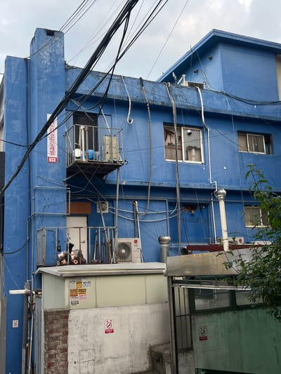 A multi-story blue building, located in Gangseo-gu, South Korea, is depicted under an overcast sky. The building's facade is extensively covered with exposed black electrical wires, white and grey utility conduits, and piping. On the upper-left, a small metal-railed balcony extends from a doorway, containing a white cylindrical object and blue textile. Above this balcony, a vertical white sign with Korean text reads "마스터코인노래방" (Master Coin Karaoke) and "3층" (3rd floor). A lower metal-railed balcony is present beneath the upper one, adjacent to a white door, and holds indistinct dark objects. An outdoor LG air conditioner unit is positioned on a flat, light-colored roof section below the main blue structure. Several cylindrical metal vents protrude from the building and the lower roof section. Multiple rectangular windows are visible across the blue facade; one window on the second visible floor exhibits a broken pane with missing glass. The base of the building transitions to a lighter-colored wall with brick detailing on the left. A red "No Smoking" (금연) sign is affixed to this lower wall. To the right, a partially visible green metal gate also displays a "No Smoking" sign. Green foliage is visible on the far right. No individuals or active interactions are present in the scene.