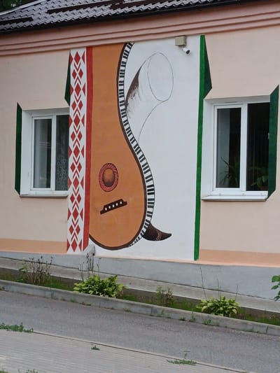This is an outdoor, eye-level, medium shot of a building mural. The mural, painted on a light peach colored wall, depicts a large brown lute with a keyboard pattern along its neck. To the left of the lute is a white column with a red and white geometric pattern. A green vertical stripe runs along the right edge of the mural. Two white-framed windows are visible, one on each side of the mural. The left window has a sheer curtain visible inside, while the right window shows potted plants. A security camera is mounted on the wall above the mural to the left of the lute's neck. The building has a dark-tiled roof. In front of the building, a gray paved sidewalk runs parallel to the street. A concrete curb separates the sidewalk from a grassy area with small plants. The street is paved with asphalt. The scene appears to be in a public area, possibly a street or a pathway. The location is identified as Смілавіцкі сельскі Савет, Belarus.