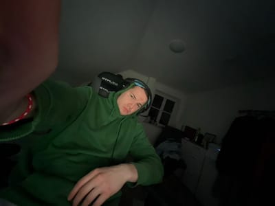 The image displays an indoor room in Benningen, Germany, under low illumination. A young adult male occupies the foreground, facing the camera. He is wearing a plain green hooded sweatshirt and a blue-green over-ear headset. A red and white braided bracelet is visible on his left wrist. His left arm is partially visible, extending towards the camera, indicative of a self-portrait. The male exhibits a neutral facial expression. Behind him, a black gaming chair featuring "GTPLAY" branding is discernible. The room's background includes light-colored walls, a ceiling-mounted circular light fixture, and a multi-paned window. Below the window, a white dresser or cabinet is present, holding several small, indistinct objects. Light-colored textiles are observed on surfaces within the room, and a dark, vertical object is partially visible on the far right.