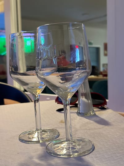 The image shows two wine glasses on a table, with reflections of a brightly lit environment, possibly a restaurant or bar, visible in them. There are also a salt shaker and a pair of red-rimmed sunglasses on the table. The scene appears to be indoors, with a blurry background suggesting other tables and patrons. The overall lighting is dim, with colorful light sources creating reflections in the glasses. The image is taken from a close-up perspective, focusing on the glasses and the immediate table setting. It is not possible to determine the exact location in El Escorial, Spain, from the visual information provided.