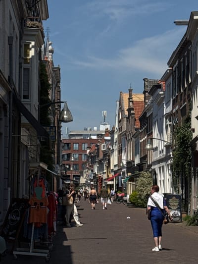 The image captures a street scene in the city of Deventer, Netherlands. The street is lined with traditional Dutch architecture, featuring narrow, multi-story buildings with characteristic gabled roofs. The buildings display a mix of architectural styles, with some having brick facades and others painted. Shopfronts are visible along the street, with awnings and displays of goods, including clothing racks with T-shirts and other apparel.
Several people are present on the street. In the foreground, a woman with short brown hair is walking away from the camera, wearing a blue top and dark pants. She has a bag slung over her shoulder. To the left, near a rack of clothes, a person is standing and possibly interacting with the clothing. Further down the street, other individuals are walking, some appear to be browsing shops. One person in the mid-ground is wearing a backpack and looking at their phone. In the background, more people are visible, and the street extends into the distance, with more buildings and shops.
The street surface is paved. A streetlamp with a distinctive glass shade is mounted on a curved arm extending from a building. The sky is clear with some wispy clouds. The sun is casting shadows on the street, indicating it is daytime. The overall atmosphere suggests a lively commercial area.