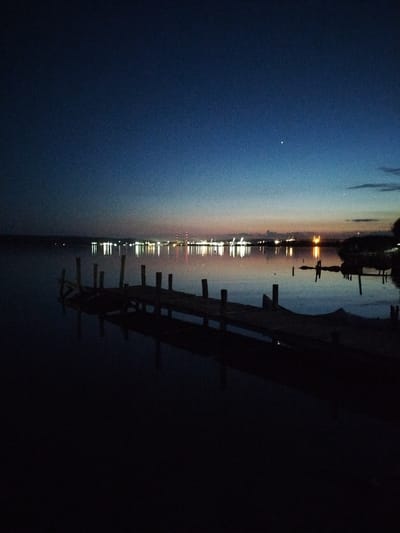 The image depicts a twilight scene at a dock on the Black Sea near the town of Kazashko, Bulgaria. The sky transitions from a deep navy blue at the top to a lighter, soft gradient of blues and pinks near the horizon. A small, crescent moon is visible in the upper portion of the sky, along with a faint star.
In the foreground, a dark, wooden pier extends into the calm water. The pier is made of planks and supported by pilings, some of which are submerged. The water reflects the dim light of the sky and the distant lights.
In the background, across the water, a cityscape is illuminated with numerous lights. These lights appear to be from buildings and possibly industrial structures, with some taller structures featuring vertical red and white lights, suggesting they might be communication towers or industrial smokestacks. The lights create a shimmering reflection on the water's surface. The overall atmosphere is serene and peaceful, capturing the transition from day to night.