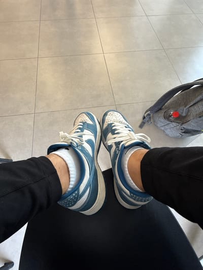 A person's legs are shown from the knees down, sitting in a chair. They are wearing black pants and white socks. The person's feet are wearing a pair of blue and white Nike sneakers. A grey backpack is sitting on the floor to the right. The floor is made of light grey tiles, and the chair is black. The image was taken in Kfar Monash, Israel.