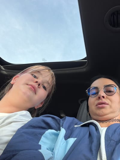 The media file shows a close-up selfie taken from a low angle inside a vehicle, looking upwards towards the sky. Two individuals are visible. In the foreground, a young girl with blonde hair, some streaks of pink, and blue eyes looks directly at the camera with a neutral expression. She is wearing a white t-shirt. Behind her and to the right, an adult woman with dark hair and glasses is also looking towards the camera. She is wearing a blue and light blue jacket and has a choker necklace on. Her expression appears serious. The background shows a sunroof with the sky visible through it, suggesting it is daytime. The scene is likely inside a car with a dark interior, indicated by the presence of a car vent. There are no visible landmarks or specific details that definitively pinpoint the location to Шолохово, Russia, though this context is provided. The weather appears to be clear and bright due to the visible sky.