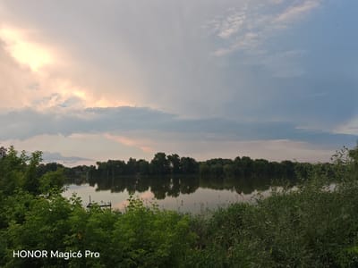 This is a landscape photograph taken in Turbiv, Ukraine. The scene depicts a tranquil lake surrounded by lush greenery and a dense line of trees on the opposite shore. The water is calm, reflecting the trees and the sky. In the foreground, dense foliage and tall reeds frame the view of the lake. A few small, dark objects, possibly boats or buoys, are visible on the water.
The sky is a prominent feature, with dramatic clouds that suggest a transition in weather or time of day. The left side of the sky is illuminated by a bright, diffused light, possibly from the setting or rising sun, casting warm, pastel tones of yellow and pink onto the clouds. The rest of the sky is covered by a softer, grayish-blue cloud cover. A few very small, dark specks, likely birds or insects, are scattered across the sky.
The time of day appears to be either early morning or late evening, given the soft lighting and the atmospheric conditions in the sky. The weather seems to be mild, with a peaceful and serene mood conveyed by the still water and the natural landscape.
In the bottom left corner of the image, the text "HONOR Magic6 Pro" is visible, indicating the device used to capture the photograph. No people are visible in the image. The overall impression is one of natural beauty and a quiet, undisturbed environment.