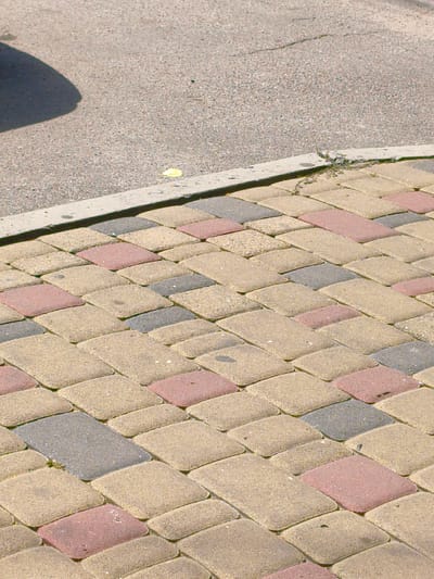 The image shows a close-up, angled view of a paved sidewalk made of interlocking pavers in a herringbone pattern. The pavers are of three colors: beige, pinkish-red, and gray. The sidewalk is bordered by a concrete curb. Behind the curb, a section of asphalt pavement is visible, with a shadow of an unseen object casting across it from the left. The asphalt appears to have a crack running through it. A small yellow leaf or piece of debris is visible on the asphalt near the curb. The scene is outdoors and appears to be a street or walkway. The location is identified as Velyka Bahachka, Ukraine.