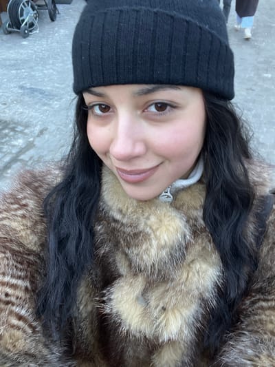 The image presents a close-up, selfie-style portrait of a young woman situated outdoors in Gstaad, Switzerland. She possesses dark, wavy hair and brown eyes, looking directly at the camera with a subtle smile. Her attire consists of a black knit beanie and a multi-toned light brown and dark brown patterned fur coat, beneath which a light-colored collar with a metallic zipper pull is visible. The immediate background features a grey, paved surface, indicative of a street or pedestrian area, appearing potentially damp or icy. In the upper left background, a wheeled utility cart equipped with a hose reel or cable spool is partially visible. Further in the upper right background, blurred figures of several individuals are discernible, suggesting a public thoroughfare with pedestrian activity.