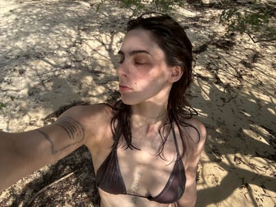 The image features a selfie of an individual, appearing female, reclining on sandy ground in Baan Nai Wok, Thailand. The subject has dark, wet hair, closed eyes, and fair skin displaying freckles and flushed cheeks. She is attired in a dark-colored bikini top. Distinct tattoos include a geometric or tribal pattern on the upper right arm and three vertical characters on the sternum. The immediate environment consists of light-colored sand, marked by scattered dry leaves and organic debris. Bright sunlight casts dappled shadows from overhead foliage across the sandy surface.