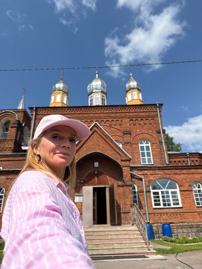 A blonde woman wearing a pink baseball cap and a pink and white striped shirt is taking a selfie outdoors. She is looking at the camera and smiling slightly. Behind her, a red brick building with a yellow-domed roof and multiple small windows is visible. The building appears to be a church or a religious institution. The sky above is blue with some scattered white clouds. The woman is positioned in the foreground, with the building in the background. The scene suggests a sunny day in Chudovo, Russia.