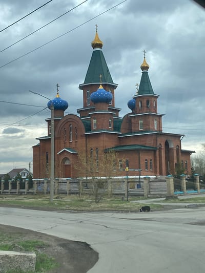 The image depicts a Russian Orthodox church located in Usolye-Sibirskoye, Russia. The church is constructed primarily of red brick with distinctive green roofs and onion-shaped blue domes adorned with gold stars. The building features multiple levels and a variety of architectural details, including arched windows, decorative moldings, and crosses atop the domes. In the foreground, there is a paved road with cracks and patches. Utility poles with overhead wires are visible on the left side of the image. The sky is overcast with grey clouds. To the side of the church there are fences and greenery, suggesting the proximity of a residential area. A dog is observed in the middle of the road.