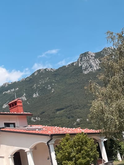 The image shows a view of the village of Budanje, Slovenia, with a house in the foreground and a mountain in the background. The house has a red-tiled roof and a red brick chimney. There is a tree to the right of the house, and the mountain is covered in greenery with some rocky outcrops. The sky is blue with a few scattered clouds.