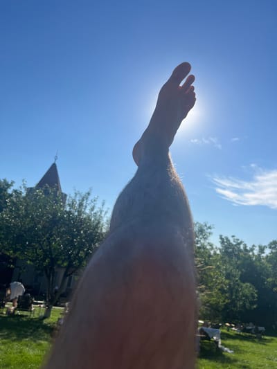 The image, taken in Stăuceni, Moldova, is a close-up, low-angle shot of a person's leg and foot against a bright blue sky with scattered white clouds. The sun is visible, creating a lens flare and silhouetting the foot. The foot is bare, with toes clearly defined, and appears to be raised. The leg is hairy and tanned, indicating it is likely daytime and the person is outdoors.
In the background, out of focus, are trees with lush green foliage, suggesting a summer day. A building with a distinctive peaked roof and a small spire or weather vane is visible among the trees, along with hints of lawn furniture and a grassy area. The overall impression is one of relaxation and enjoyment of a sunny day, possibly in a backyard or park setting. The bright sunlight and clear sky suggest good weather, likely midday or afternoon. There is no visible text in the image.