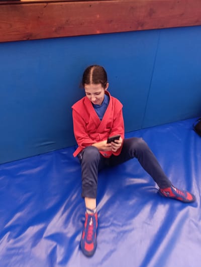 A young girl, likely under 18, wearing a red Sambo uniform over a blue t-shirt and dark pants, sits on a blue mat. Her hair is pulled back in a braid. She is holding a smartphone in her hands and looking down at it. The mat is in what appears to be a Sambo training facility. The background shows a wooden beam above a blue padded wall. The location is the city of Ladyzhyn, Ukraine.