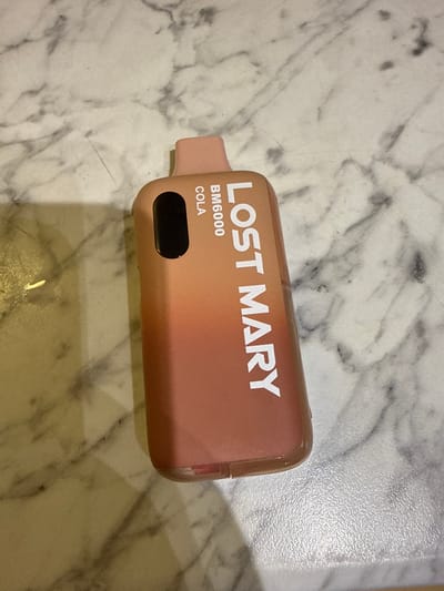 The image displays a single Lost Mary BM6000 Cola disposable vape device, positioned in Grays, United Kingdom. The device is angled on a light-colored surface characterized by a grey and white marble pattern. The vape unit features an ombre design, transitioning from a light peach tone at the integrated mouthpiece and upper section to a darker reddish-brown hue at its base. Prominent white text "LOST MARY" is displayed along the side of the device, with "BM6000 COLA" printed vertically near the top. An opaque black, elongated oval feature is present on the upper part of the device. A transparent lower section of the vape reveals a red internal component. No people, subjects, actions, or interactions are visible in the scene.