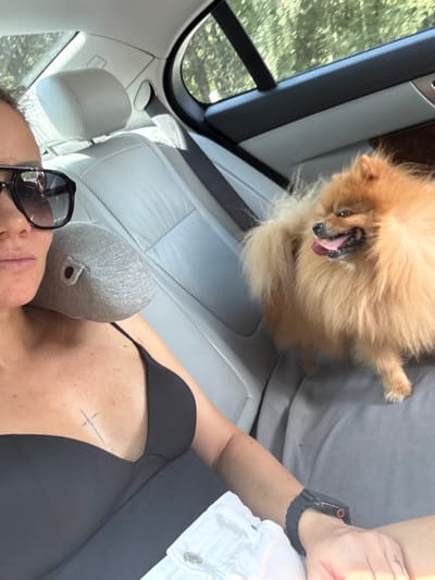 A medium shot shows a woman in the back seat of a car, wearing sunglasses and a black top. She has a small grey pillow behind her head. A fluffy orange Pomeranian dog is sitting next to her, panting with its tongue out. The interior of the car is visible, with light-colored leather seats and black trim. Outside the car windows, trees and foliage can be seen, suggesting a suburban or rural setting. The image does not provide any information to indicate the location is Lüneburg, Germany.