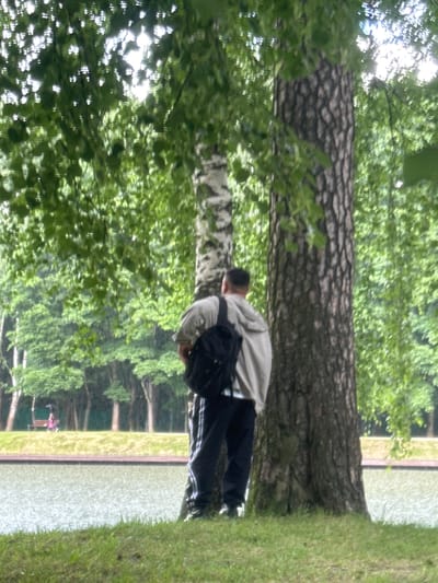 A person is seen from the back, wearing a light-colored hoodie and dark pants with white stripes. They have a black backpack on. The person is standing near the thick trunk of a tree, surrounded by lush green foliage and dappled sunlight filtering through the leaves. In the background, a body of water is visible, with a grassy bank on the right and what appears to be a paved path or edge along the water. The scene suggests a park or natural setting. The image is oriented vertically, with the sky and trees appearing at the top and bottom. There is no visible text in the image. The overall impression is a quiet, outdoor moment, possibly a pause in a walk or exploration. The lighting suggests it could be daytime, perhaps late morning or afternoon, with bright sunshine. The setting appears to be a park or garden area, possibly near a lake or pond, given the presence of the water.