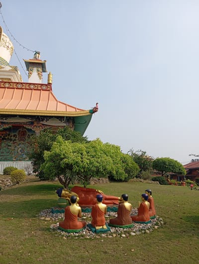 The image depicts a serene garden scene in Lumbini Sanskritik, Nepal, featuring a reclining Buddha statue surrounded by smaller monk figures. The Buddha statue is lying on its right side, adorned in orange robes, with its head painted a dark blue. The monk statues, also in orange robes, are seated in a circle around the Buddha, facing inward, suggesting a meditative or devotional setting. They all sit on a base of stones. Behind the statues is a lush green lawn with trees and shrubs. In the background, a building with intricate murals and an orange tiled roof is visible, contributing to the location's cultural and religious context. The overall impression is one of peace and reverence, typical of Buddhist sites.