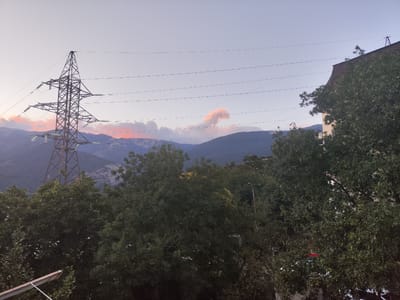 The image captures a late afternoon or early evening scene with a wide, clear sky transitioning to dusk. In the foreground, dense green foliage from trees obscures the lower portion of the frame, with some branches and leaves visible. A tall, latticed power line tower stands prominently on the left side, supporting multiple high-voltage wires that stretch across the scene. In the mid-ground, a range of dark, hazy mountains is visible, with a faint reddish-pink glow from the setting sun illuminating some of the clouds above them. To the right, the top of a multi-story building with a brown roof is partially visible, peeking through the trees. The overall atmosphere is serene, with the natural landscape of mountains and trees juxtaposed against the man-made structures of the power lines and building. The image does not contain any visible people or ongoing actions. The location is specified as Massandrovsky Village Council, Ukraine.