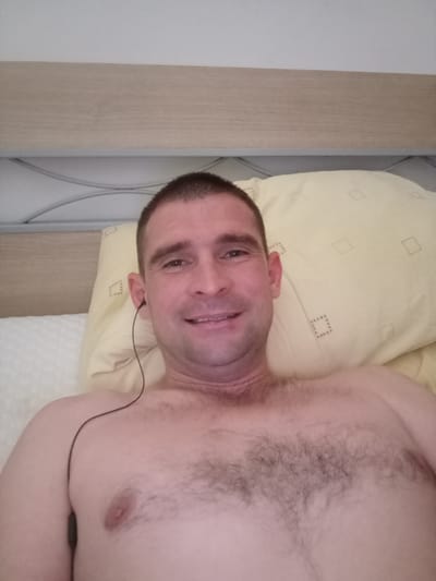 The image shows a man lying in bed, looking at the camera. He is shirtless and wearing black earbuds. The man has short brown hair and a short beard. He is smiling. Behind him is a yellow pillow with a diamond pattern. The bed is covered with a white sheet. The headboard of the bed is visible in the background. The lighting suggests it is daytime. There is no visible text in the image. The location context is Civitanova Marche, Italy, but there are no specific visual cues in the image to confirm this.