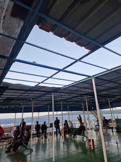 The image shows the interior of a ferry in Bulusan, Indonesia. Passengers are seated on benches under a covered section of the ferry. The roof is made of corrugated metal with blue metal supports. The ocean is visible in the background. A red fire extinguisher is also visible.