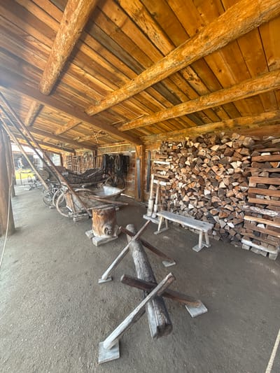 This image depicts the interior of a rustic wooden structure, likely a barn or workshop, located in Shushenskoye, Russia. The scene is filled with historical artifacts and supplies. In the foreground, a large pile of chopped firewood is stacked against the back wall. To the left, several horse-drawn carriages and carts are parked, some appearing old and weathered. A wooden bench sits in front of the firewood. Various tools and equipment, including what appear to be agricultural implements and harnesses, are visible throughout the space. The ceiling is constructed of rough-hewn wooden beams and planks, contributing to the primitive atmosphere. The floor is dirt. A person is faintly visible walking in the background through an open doorway on the left.