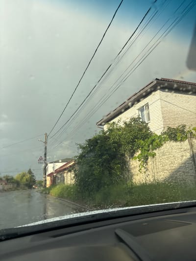 The image depicts a street scene in Pokotylivka, Ukraine, viewed through a wet windshield. On the right, a light-colored brick house with a brown roof sits behind a textured concrete fence. Lush green vegetation covers the fence and climbs the side of the house. To the left, a wet asphalt road reflects the overcast sky and overhead power lines. A street sign indicating a pedestrian crossing 100 meters ahead is visible on a utility pole, along with a streetlight. The overall atmosphere suggests recent rainfall or ongoing inclement weather, with water droplets visible on the windshield. The scene appears to be a quiet residential street.