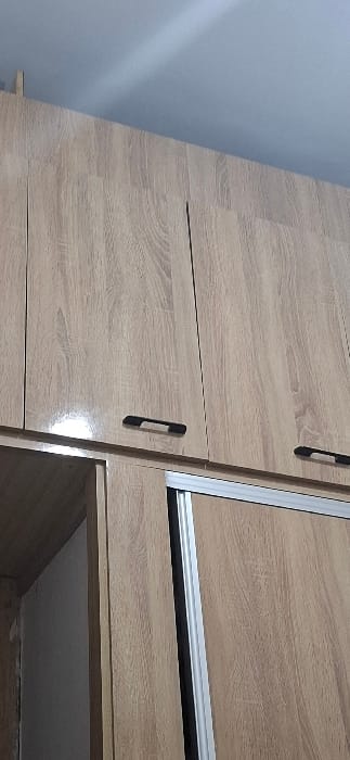 The image displays an indoor scene in Skhirat, Morocco, featuring a multi-component wooden wardrobe or storage unit. The unit is constructed from light-toned wood-grain laminate material. The upper section consists of three overhead cabinet doors, with the two central and right doors fully visible, each equipped with a black horizontal pull handle. These upper door surfaces exhibit a glossy finish, reflecting an overhead light source. Directly below the upper cabinets, the unit includes a sliding door section, characterized by silver-colored metal tracks along its edges, notably a vertical track on the left side of the sliding panel. To the immediate left of the sliding door, an open compartment or partially open door reveals a light-colored interior surface. Above the entire unit, a plain light blue ceiling is visible, displaying a subtle diagonal highlight or shadow across its surface. No individuals, actions, or interactions are present.
