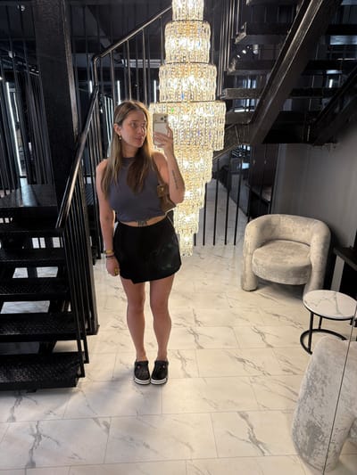 In the image, a woman stands on a marble-tiled floor, taking a selfie with her phone. She is wearing a grey cropped tank top, a black skirt, and black clogs. A brown shoulder bag is slung over her left shoulder. To her left is a black metal staircase with industrial-style steps and railings. To her right, a plush, light-colored armchair sits next to a small round table with a marble-patterned top. A large, multi-tiered crystal chandelier hangs prominently in the background, illuminating the scene. The overall aesthetic suggests an indoor, likely commercial or residential, space with a modern and luxurious design. There is no information to suggest the location is in Укромновское сельское поселение, Ukraine.