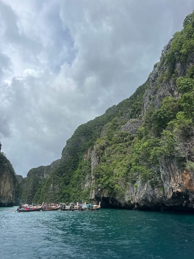 The image depicts a body of water with numerous longtail boats moored together. These boats are characteristic of Thailand, particularly in popular tourist areas. The water is a vibrant turquoise color, reflecting the overcast sky. Towering, lush green limestone cliffs rise steeply from the water on either side of the frame, creating a dramatic and natural landscape. The cliffs are covered in dense vegetation, with trees and foliage clinging to the rocky surfaces. The sky is cloudy, with varying shades of gray and white, suggesting a potentially overcast or approaching weather system. The scene appears to be a bay or cove, typical of the island scenery found in southern Thailand. The specific location is Haad Yao, Thailand, a coastal area known for its beautiful beaches and limestone karst formations. The boats are likely used for tours or transportation in this scenic region.