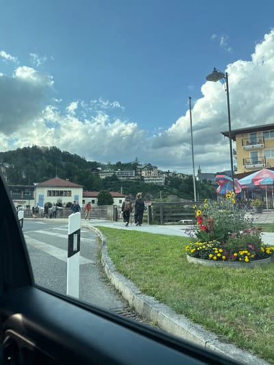 The image depicts a street scene in Berchtesgaden, Germany, on a partly cloudy day. In the foreground, a car's interior is visible on the left side, with the side mirror and part of the dashboard in frame. A pedestrian crossing with a black and white striped pole is in the middle ground, leading to a sidewalk.
Several individuals are walking on the sidewalk. One person in the foreground is seen from behind, wearing a backpack and dark clothing, walking away from the camera. Another person, also from behind, is dressed in dark clothing and carrying a backpack. Further down the sidewalk, a group of people are walking and standing near a building with a red roof and beige facade.
Beyond the sidewalk, there's a fenced area with a grassy embankment. A brightly colored flower bed filled with yellow, red, and purple flowers is situated on the grass. To the right, a yellow building with balconies stands prominently. Several tables and chairs are set up outside this building, some covered by red and blue umbrellas. A lamppost is visible near the yellow building, and a taller, thinner pole stands beside it.
In the background, a hillside covered with lush green trees is dotted with various buildings, including several larger, multi-story structures that appear to be hotels or apartments. A prominent church steeple rises above the trees in the distance. The sky is a vibrant blue with scattered white clouds, some of which are large and fluffy. The overall impression is of a bustling town square or pedestrian area within the scenic landscape of Berchtesgaden.