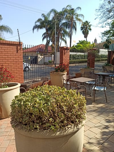 The image depicts an outdoor seating area in Pretoria, South Africa. In the foreground, there are two large concrete planters, one on the left containing red flowers and another on the right filled with a dense green shrub. A metal barbecue grill sits in front of the brick wall to the left. Behind the grill, a black metal fence separates the seating area from the street. A white car is visible parked on the other side of the fence. The seating area itself features several metal tables and chairs, some with blue patterned cushions. In the background, palm trees are prominent, and the silhouette of a building with a dome can be seen in the distance. The ground is paved with interlocking bricks. The sky is clear and blue. The overall scene suggests a restaurant or cafe setting.