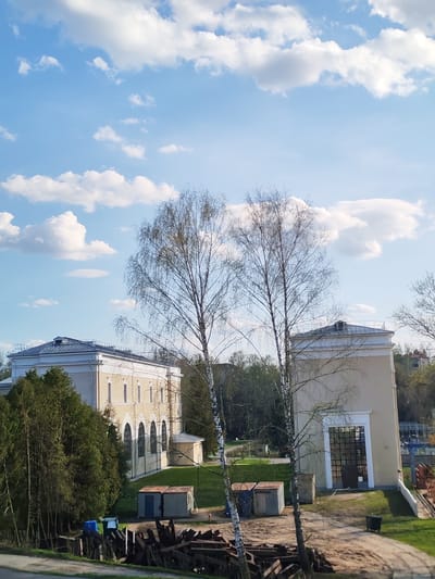The image depicts a daytime scene in what appears to be a suburban or semi-rural area of Водник Снт, Russia. The sky is blue with scattered white clouds. Two buildings stand prominently in the midground. The building to the left is a two-story structure with arched windows and a light-colored facade. To the right is a taller, narrower building with a large glass door. Between the two buildings is a grassy area with several smaller structures or sheds. Two bare trees are positioned in the foreground, partially obscuring the view of the buildings. In the very foreground, there's what seems to be a pile of debris or construction materials. The overall impression is of a quiet, somewhat industrial or utility-focused section of the city.