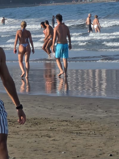 At the beach in Playa Del Inglés, Spain, several individuals are enjoying the water and sand. In the foreground, a person from the chest down is partially visible, wearing striped shorts and a black wristband. To their right, a woman with blonde hair is wearing a blue bikini and walking towards the water. Behind her, two individuals, a man and a woman, are standing in the ocean, with the woman wearing a pink bikini. Further back, a man in blue swim trunks is walking out of the water. To his left and slightly further back, a man with gray hair and a woman in a white bikini are standing in the ocean. In the distance, other beachgoers are visible, as well as beach umbrellas and the ocean with waves. The sand is wet near the water's edge and reflective, showing the ripples from the waves.