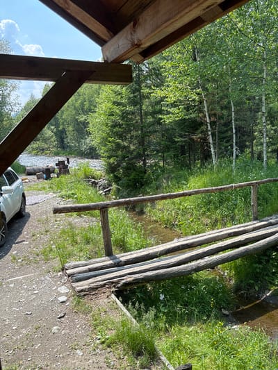 This is an outdoor, eye-level, vertical shot captured on a sunny day in Bol'shaya Rechka, Russia. The scene features a rustic wooden bridge constructed from logs that spans a small, shallow stream. The bridge is in the foreground and is weathered, with visible grain and imperfections in the wood. To the left of the bridge, there is a gravel path and a white SUV parked. Beyond the stream and bridge, dense green foliage from trees and undergrowth creates a natural, wooded environment. A river with flowing water is visible in the background, surrounded by more trees. The sky above is blue with scattered white clouds. The overall impression is one of a remote, natural setting, possibly a campsite or a recreational area by a river. There are no people visible in the image.