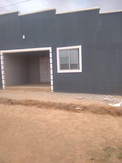 The image depicts the exterior of a newly constructed, dark gray, single-story building in Goni Gara, Nigeria. The building has a light-colored trim along the roofline, which has a stepped or crenellated design. A single, white, outdoor light fixture is mounted on the front wall.
The front of the building features a wide, open entrance or porch area, bordered by a white frame. Inside this opening, a metal door is visible. To the right of the entrance, there is a window with multiple panes, framed in white. The building is set on a dirt ground, with a trench dug in front of it, suggesting ongoing construction or landscaping work. Some rubble and debris are visible near the building.
The sky is overcast, indicating a cloudy day. The time of day appears to be daytime, given the ambient light. The scene is one of a building under construction or recently completed, in a rural or semi-rural setting, as suggested by the dirt surroundings and the style of the building. There is no visible text in the image. No people are present in the frame. The overall impression is one of a functional, unfinished or newly finished structure.