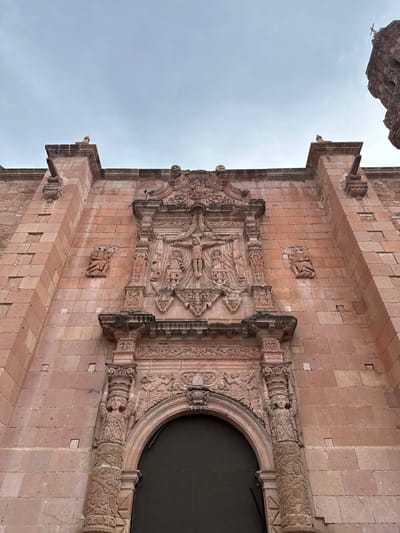 This is a low-angle exterior shot of a stone facade of a historic building, likely a church, in Zacatecas City, Mexico. The facade is constructed of large, pinkish-red sandstone blocks. The central focus is an ornate stone relief carving above an arched doorway. The relief depicts a crucifixion scene with Jesus on the cross, flanked by figures. Below this, smaller carvings include what appear to be cherubs or angels. Flanking the central relief are two pilasters, each topped with a carved figure supporting the elaborate entablature. The pilasters themselves are decorated with intricate patterns, including what seems to be a vine motif. On the left and right sides of the main facade, higher up, are two small, figural carvings in relief, appearing to depict seated figures. Above the facade, the sky is overcast with a pale blue hue. The overall impression is of weathered, detailed stonework typical of colonial architecture.