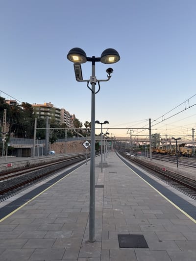 The image captures a scene at a train station platform. The primary focus is a tall, gray metal light pole with two dome-shaped lights and a security camera attached. The platform is paved with large rectangular stones and features a yellow safety line along the edges where it meets the train tracks. Tracks run on both sides of the platform.
A white sign with black lettering "110" is visible, presumably indicating a speed restriction. Buildings, trees, and hills can be seen in the background. A maintenance train with yellow and black coloring is parked on tracks to the right. Overhead electrical wires are strung across the scene.
The photograph was taken at a train station in Tarragona, Spain.