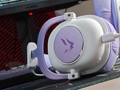 This is a close-up, eye-level shot of a white and purple gaming headset resting on a mousepad. The headset is angled, with the right earcup prominently displayed. The earcup has a circular, purple insert featuring a stylized, purple lightning bolt design. The headband, also purple, curves over the earcup. A white cable connects to the earcup. The background shows a dark computer monitor with a red indicator light and a red lattice pattern on its stand. The scene appears to be a gaming setup. No people are visible, and the time of day, weather, or specific location cues beyond Russia are not discernible. The image focuses on the details of the headset and its immediate surroundings.