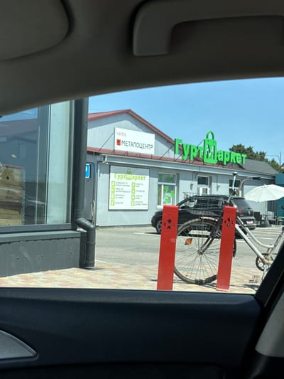 This is a photo taken from inside a vehicle, looking out at a storefront. The most prominent text visible is "ГуртМаркет" (GurtMarket) in large green letters, which appears to be the name of the store. Below this, in smaller text, are categories of products such as "М'ЯСНІ ВИРОБИ" (Meat products), "МОРЕПРОДУКТИ" (Seafood), "МОЛОЧКА ТА СИРИ" (Dairy and cheese), and "БАКАЛІЯ" (Groceries). There is also text that reads "МЕТАЛОЦЕНТР" (Metal Center), indicating another business or department.
In front of the store, a bicycle is parked. It has a basket in the front and is secured by two bright red vertical posts. The ground in front of the store is paved with a yellow and reddish-brown brick pattern. The building itself appears to be a commercial structure with a grey, corrugated metal exterior and a reddish-brown roofline. A white umbrella is visible behind the bicycle.
The sky is blue and appears clear, suggesting it is daytime and likely sunny. The overall scene suggests a commercial area or a shopping district. The photo is taken from a slightly angled perspective, with the interior of the car's dashboard and door frame visible in the foreground on the left and right, respectively. The presence of the car interior implies the photo was taken by someone in transit. The location context of Sarny, Ukraine, is provided separately.