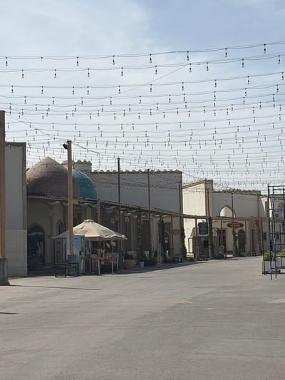 The image depicts a wide outdoor area, possibly a pedestrian plaza or street, under a clear, bright daytime sky. Hundreds of small, clear string lights are strung across the sky, creating an overhead canopy effect.
On the right side of the frame (after rotating 90 degrees clockwise), a series of low-rise buildings with light-colored facades line the area. Architectural features include arched windows and decorative elements. Among these buildings, a prominent blue-domed structure and a smaller, brown-tiled dome are visible. Several poles support the string lights and other structures.
In front of the buildings, a large, light-colored umbrella bearing the "Coca-Cola" logo is visible, alongside some outdoor furniture and display stands. The ground is a wide expanse of paved asphalt or concrete.
Notable details include:
- A brown sign on one building reads "GRAND BAZAAR".
- Below the Grand Bazaar sign, another sign partially reads "PISHMANGI RESTAURANT".
- A tall, dark rectangular sign in front of a building displays white text. The visible lines include "SHAHRISABZ", "WELCOME TO THE HEART OF THE...", "WE SERVE UZBEK PILAF SAMARKAND STYLE", and "TRADITIONAL FOOD".
- Small American flags are displayed near the tall black sign.
No people are visible, and no specific activity is taking place; the scene appears still. The bright sky indicates it is daylight. The architecture and reference to "Uzbek Pilaf Samarkand Style" align with the provided location context of Uzbekistan.
