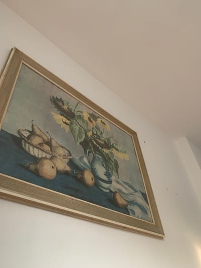 A framed still life painting hangs on a white wall. The painting depicts a basket of pears, several loose pears on a blue surface, and a vase of sunflowers. The frame is light brown and appears to be made of wood. The painting is hung at an angle, with the top edge closer to the camera. The room appears to be dimly lit, with light coming from the right side of the frame, casting shadows on the wall. There are no people visible in the image. This scene is not located in Vejprty, Czechia.